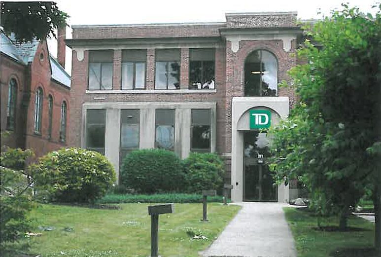 Large brick building with a green sign on the front of it, surrounded by trees, plants, grass, and a walkway